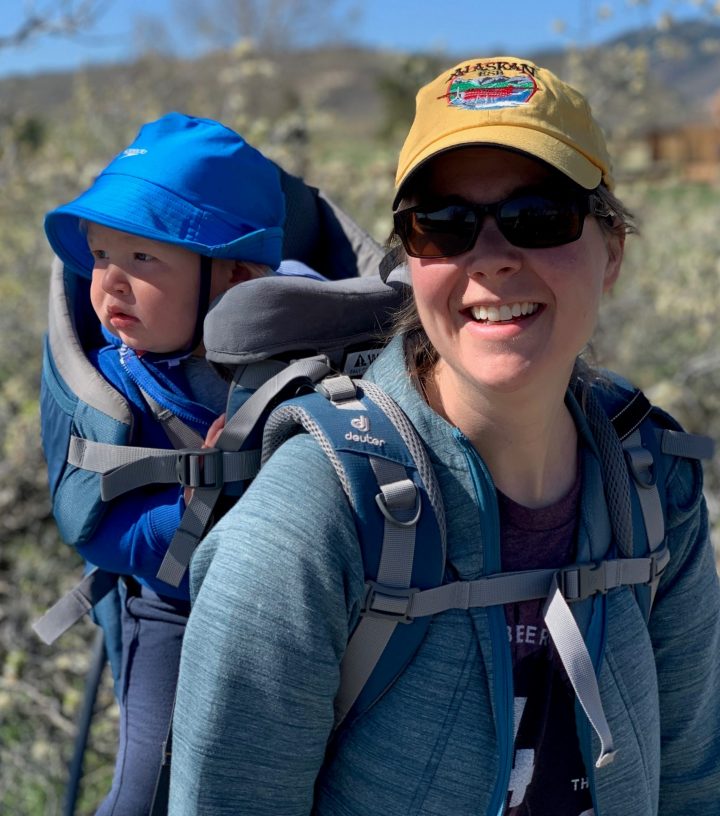 woman wearing yellow hat with baby in carrier on her back, baby is wearing blue hat